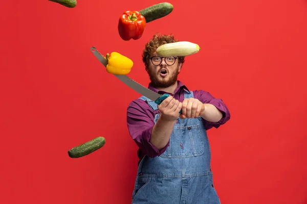 Verdure volanti. Carino uomo barbuto dai capelli rossi, contadino con coltello divertirsi isolato su sfondo rosso studio. Concetto di professione professionale, lavoro. — Foto Stock