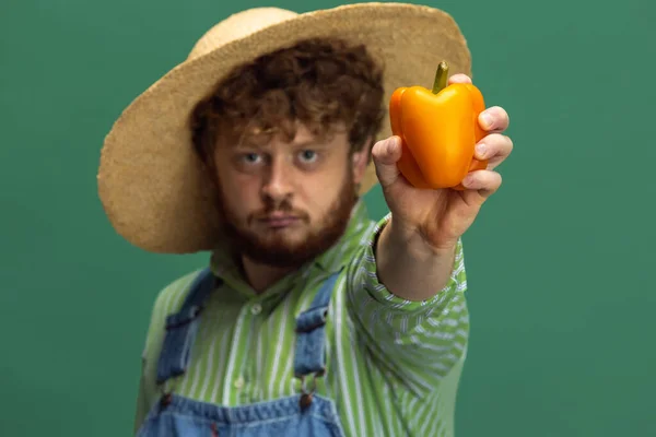 Close-up funny redheaded bearded man, farmer with yellow pepper standing isolated over green studio background. Concept of professional occupation, work. — Photo