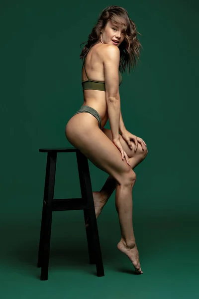 Jedna mladá atraktivní dívka, modelka v zeleném spodním prádle, plavky sedí na vysoké židli izolované přes tmavě zelené pozadí studia. Přírodní krása — Stock fotografie