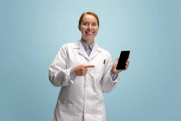 Portrait of female doctor, therapeutic or medical advisor with smartphone screen isolated on blue background. Funny happy emotions. Concept of healthcare, care medicine and humor —  Fotos de Stock