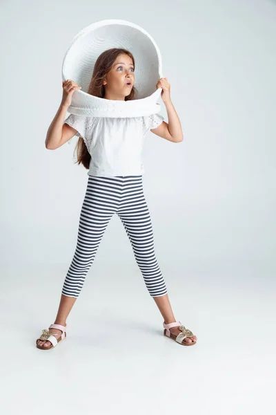 Full-length portrait of of beautiful preschool girl in big white astronaut helmet isolated on gray studio background. Childhood, education, emotion concept