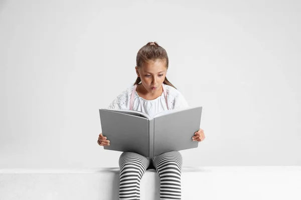 Portret uroczej białej dziewczyny, nastolatka siedząca i czytająca dużą książkę, magazyn odizolowany na szarym tle studia. Dzieciństwo, edukacja, koncepcja emocji. — Zdjęcie stockowe