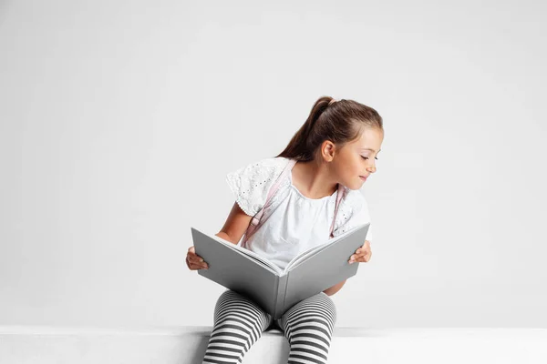 Ritratto di ragazza caucasica carina, adolescente seduto con grande libro, rivista isolata su sfondo grigio studio. Infanzia, educazione, concetto di emozione. — Foto Stock