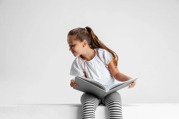 Ritratto di ragazza caucasica carina, adolescente seduto con grande libro, rivista isolata su sfondo grigio studio. Infanzia, educazione, concetto di emozione. — Foto Stock