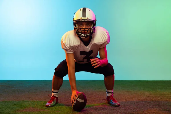 Jovem, jogador de futebol americano masculino treinando isolado em fundo de estúdio azul em luz de néon. Conceito de desporto, competição — Fotografia de Stock
