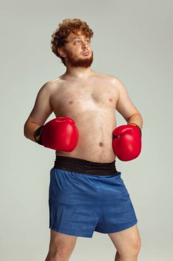 Mavi şortlu ve eldivenli komik kızıl saçlı adam gri stüdyo arka planında izole edilmiş. Spor anlayışı, mizah ve beden pozitif.