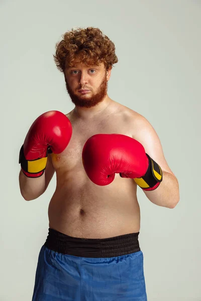 Divertente uomo dalla testa rossa in pantaloncini da boxe blu e guanti isolati su sfondo grigio studio. Concetto di sport, umorismo e corpo positivo. — Foto Stock