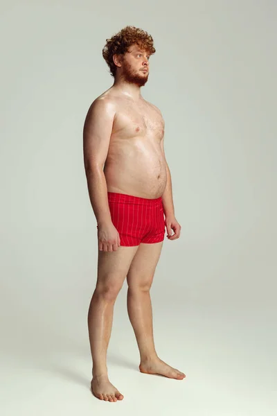 Netter rothaariger Mann in roten Badehosen, der isoliert auf grauem Studiohintergrund posiert. Konzept von Sport, Humor und Körper positiv. — Stockfoto