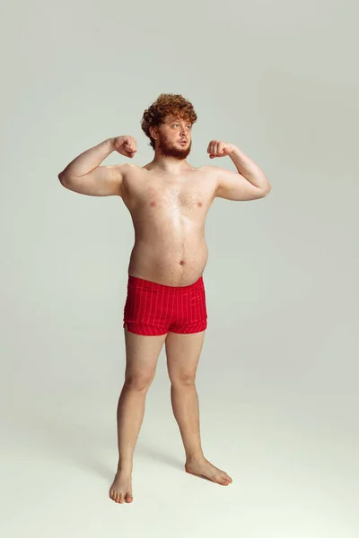 Leuke roodharige man in rode zwembroek die geïsoleerd poseert op een grijze studio achtergrond. Concept van sport, humor en lichaam positief. — Stockfoto
