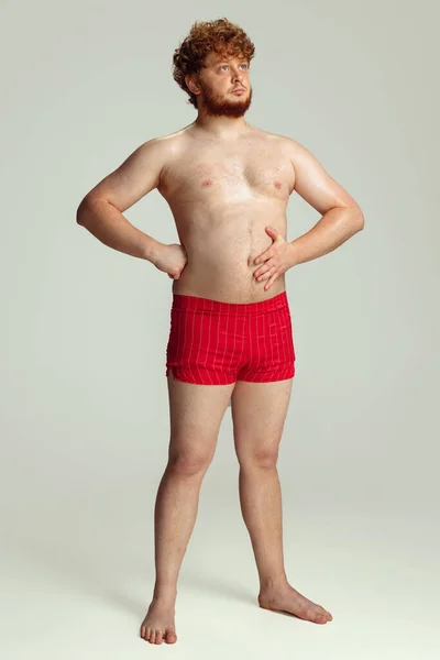 Mignon homme roux en short de bain rouge posant isolé sur fond de studio gris. Concept de sport, humour et corps positif. — Photo