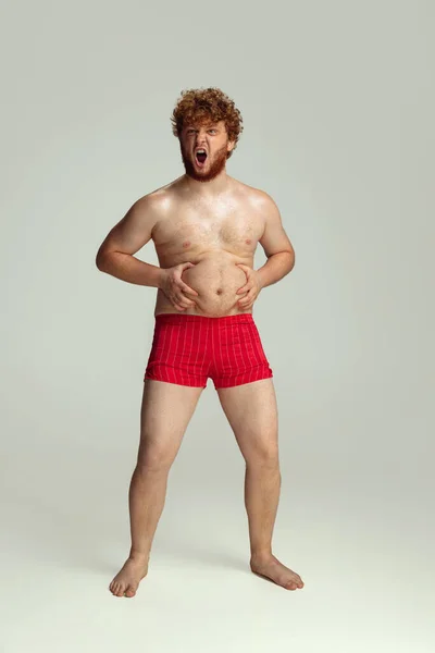 Lindo hombre pelirrojo en pantalones cortos de natación rojos posando aislado sobre fondo gris estudio. Concepto de deporte, humor y cuerpo positivo. — Foto de Stock