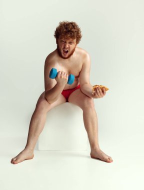 Kırmızı şortlu, gri stüdyo arka planında yalnız poz veren sevimli kızıl saçlı adam. Spor anlayışı, mizah ve beden pozitif.