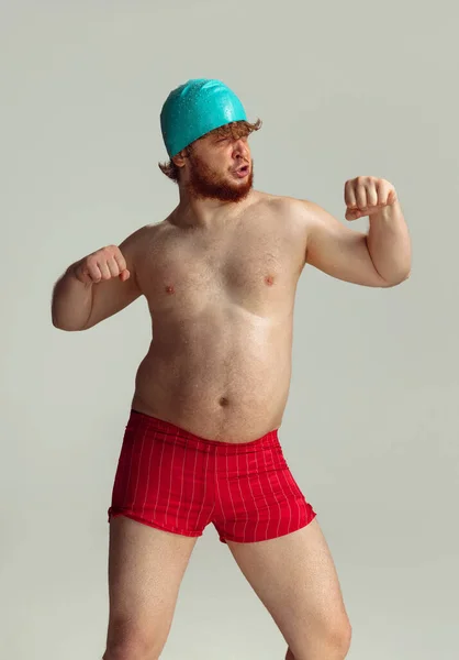 Nadador alegre engraçado. Homem ruivo bonito em shorts de natação vermelho posando isolado no fundo do estúdio cinza. Conceito de esporte, humor e corpo positivo. — Fotografia de Stock