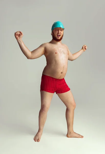 Amusant nageur joyeux. Mignon homme roux en short de bain rouge posant isolé sur fond de studio gris. Concept de sport, humour et corps positif. — Photo