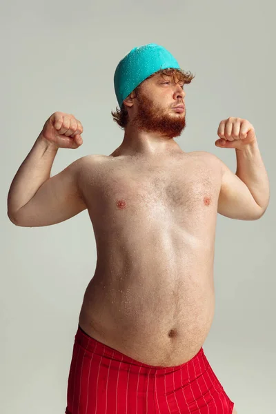 Lustige fröhliche Schwimmerin. Netter rothaariger Mann in roten Badehosen, der isoliert auf grauem Studiohintergrund posiert. Konzept von Sport, Humor und Körper positiv. — Stockfoto