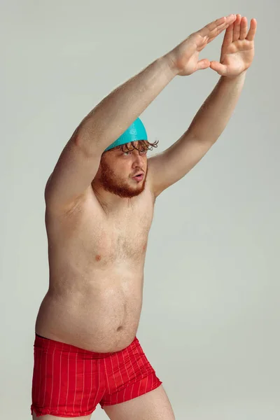 Lustige fröhliche Schwimmerin. Netter rothaariger Mann in roten Badehosen, der isoliert auf grauem Studiohintergrund posiert. Konzept von Sport, Humor und Körper positiv. — Stockfoto