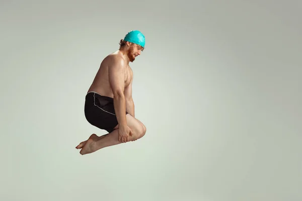 Mergulho engraçado. Homem ruivo bonito em shorts de natação vermelho posando isolado no fundo do estúdio cinza. Conceito de esporte, humor e corpo positivo. — Fotografia de Stock
