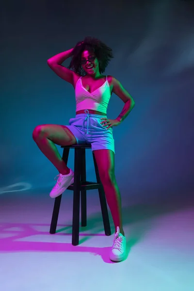 Retrato de menina africana posando isolado no fundo do estúdio azul escuro em luz de néon rosa. Conceito de emoções humanas, expressão facial, juventude, vendas, anúncio. — Fotografia de Stock