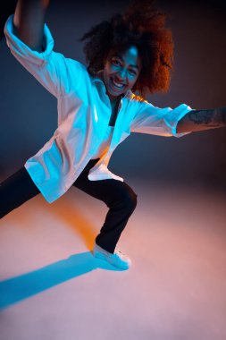 Afrikalı genç bir kızın tam boy portresi günlük kıyafetler, koyu mavi stüdyo arka planında pembe neon ışıkta izole edilmiş bir kıyafet. İnsan duyguları kavramı, yüz ifadesi, gençlik, satış, reklam.