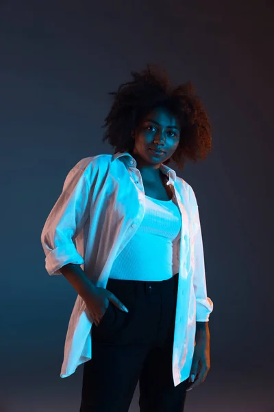 Full-length portrait of African young girl casual clothes, outfit isolated on dark blue studio background in pink neon light. Concept of human emotions, facial expression, youth, sales, ad.