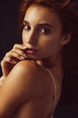 Genç, ince, bronzlaşmış, kızıl saçlı, iç çamaşırlı bir kadın. Karanlık stüdyo arka planında yalnız poz veriyor. Doğal güzellik konsepti.
