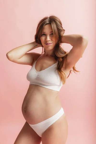 Feliz tempo, à espera. Retrato de mulher grávida bonita sorridente em lingerie isolada sobre fundo de estúdio rosa. Beleza natural, maternidade feliz, conceito de feminilidade. — Fotografia de Stock