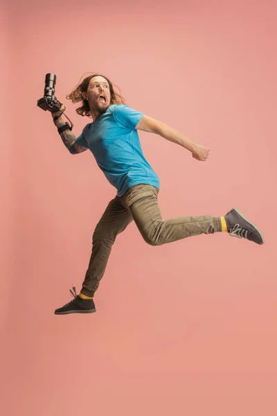 Портрет молодого чоловіка, фотографа, оператора з камерою, що стрибає ізольовано на рожевому студійному фоні. Концепція занять, роботи, смішні мем емоції — стокове фото