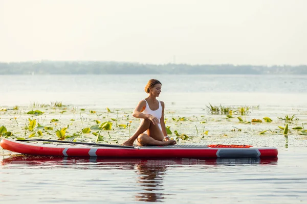 Ein junges kaukasisches schönes schlankes Mädchen sonnt sich an einem Sommerabend auf dem Fluss auf einem Paddelbrett, SUP allein. Aktives Leben, Sport, Freizeitkonzept — Stockfoto