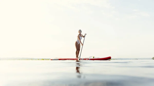 Ein junges kaukasisches schönes schlankes Mädchen sonnt sich an einem Sommerabend auf dem Fluss auf einem Paddelbrett, SUP allein. Aktives Leben, Sport, Freizeitkonzept — Stockfoto