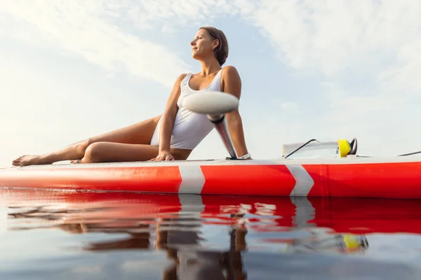 Ein junges kaukasisches schönes schlankes Mädchen sonnt sich an einem Sommerabend auf dem Fluss auf einem Paddelbrett, SUP allein. Aktives Leben, Sport, Freizeitkonzept — Stockfoto