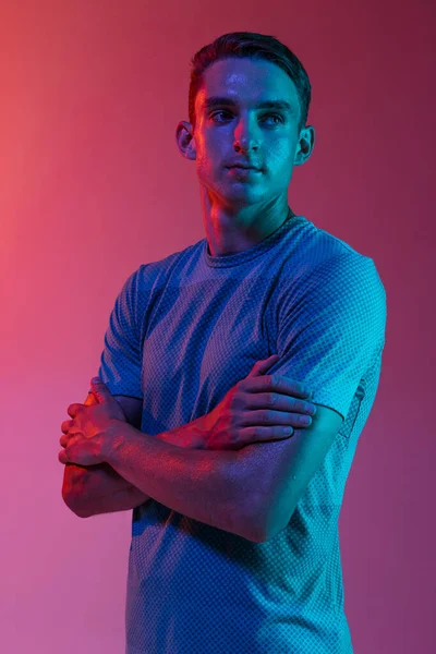 身穿运动服的白种人年轻人的肖像，站在粉红色的工作室背景上，带有蓝色的霓虹灯滤光片。肌肉男模. — 图库照片