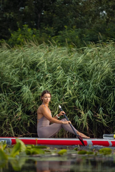 Una joven caucásica hermosa chica delgada tomar el sol en la noche de verano en el río en el tablero de paddle, SUP solo. Vida activa, deporte, concepto de actividad de ocio —  Fotos de Stock