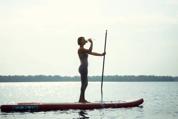 Ein junges kaukasisches schönes schlankes Mädchen sonnt sich an einem Sommerabend auf dem Fluss auf einem Paddelbrett, SUP allein. Aktives Leben, Sport, Freizeitkonzept — Stockfoto