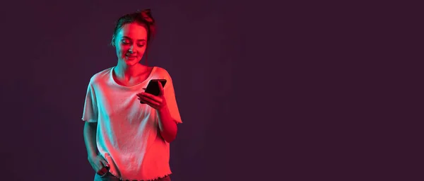 Portret de fată destul de romantic cu telefon, gadget-uri izolate pe fundal studio violet închis în lumină neon roșu, filtru. Conceptul de emoții umane, expresie facială . — Fotografie, imagine de stoc