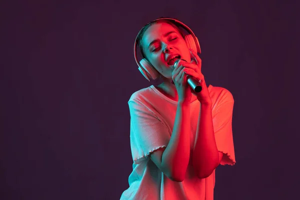 Gelukkig meisje met microfoon geïsoleerd op donker paarse studio achtergrond in rood neon licht, filter. Concept van menselijke emoties, gezichtsuitdrukking. — Stockfoto