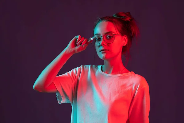 Retrato de menina bonita em roupas casuais isolado em fundo estúdio roxo escuro em luz de néon vermelho, filtro. Conceito de emoções humanas, expressão facial. — Fotografia de Stock