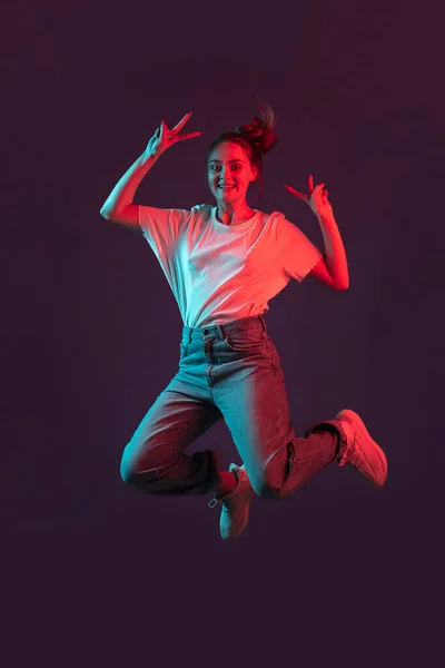 Pionowy portret pięknej dziewczyny w ubraniach casual skoków izolowanych na ciemnym fioletowym tle studio w czerwonym świetle neonowym, filtr. Pojęcie ludzkich emocji, kultury młodzieżowej — Zdjęcie stockowe