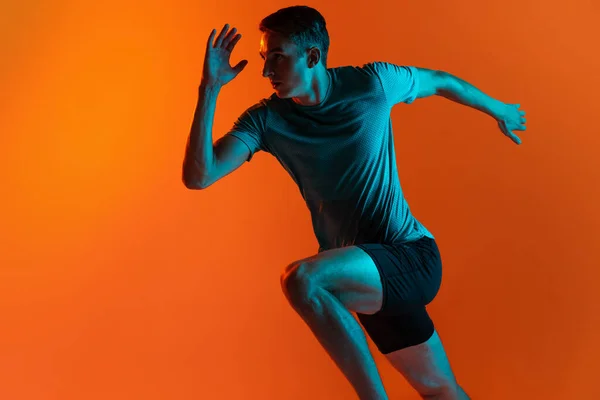 Portrat atlet pria profesional Kaukasia, pelatihan pelari terisolasi di latar belakang studio oranye dengan filter neon biru, cahaya. Pria berotot dan sportif. — Stok Foto