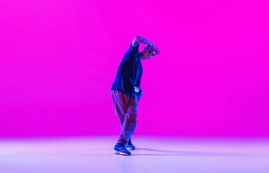 Genç stil sahibi bir adam, modern kıyafetlerle dans eden hip-hop dansçısı. Parlak mor arka planda, neon ışıklı dans salonunda izole edilmiş..