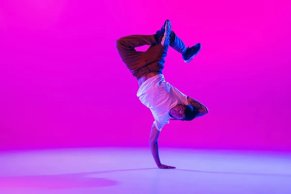 Młody stylowy mężczyzna, tancerz hip-hopowy tańczący solo w nowoczesnych ubraniach odizolowanych na jasnym tle magenta w sali tanecznej w neonowym świetle. — Zdjęcie stockowe