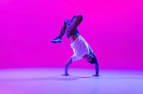 Jonge stijlvolle man, breakdance danser oefenen solo in moderne kleren geïsoleerd over heldere magenta achtergrond in danszaal in neon licht. — Stockfoto