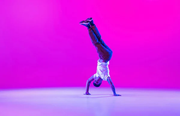 Jovem homem elegante, dançarino de hip-hop dançando sozinho em roupas modernas isoladas sobre fundo magenta brilhante no salão de dança em luz de néon. — Fotografia de Stock