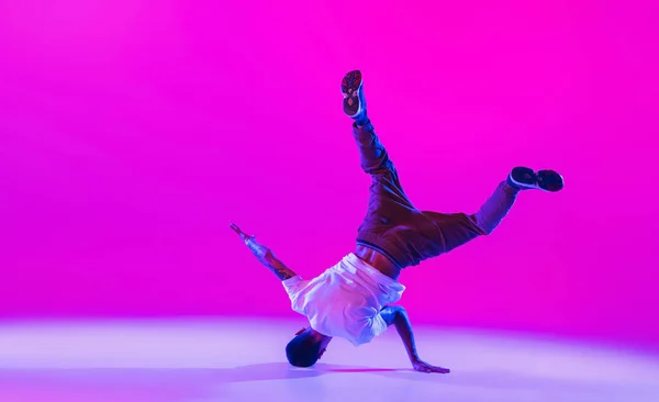 Jonge stijlvolle man, hip-hop danser dansen solo in moderne kleding geïsoleerd over heldere magenta achtergrond in danszaal in neon licht. — Stockfoto
