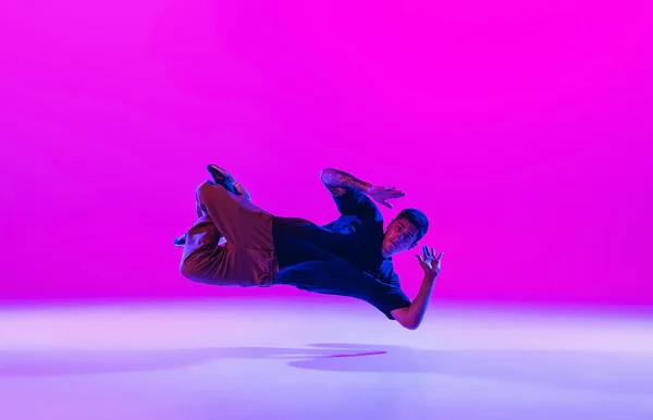 Jonge stijlvolle man, breakdance danser oefenen solo in moderne kleren geïsoleerd over heldere magenta achtergrond in danszaal in neon licht. — Stockfoto