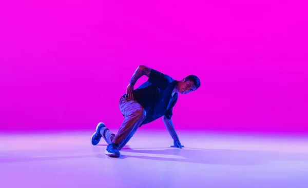 Jonge stijlvolle man, hip-hop danser dansen solo in moderne kleding geïsoleerd over heldere magenta achtergrond in danszaal in neon licht. — Stockfoto