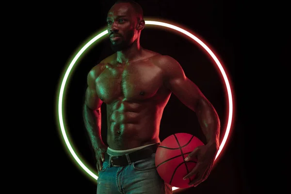 Diseño abstracto. Joven africano, atleta musculocon balón de baloncesto aislado sobre fondo negro con círculo geométrico luminiscente de neón —  Fotos de Stock