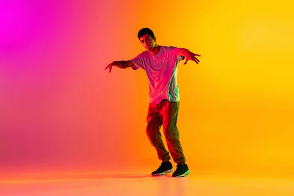 Sólové vystoupení. Mladý muž, break dance tanečnice ptacticing v ležérní oblečení izolované přes gradient růžové žluté pozadí v tanečním sále v neonovém světle. — Stock fotografie