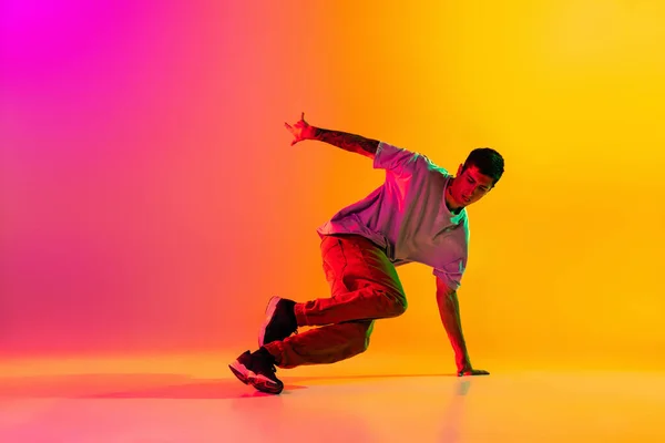 Portret van jonge stijlvolle man, breakdance danseres training in casual kleding geïsoleerd over hellend roze gele achtergrond in danszaal in neon licht. — Stockfoto