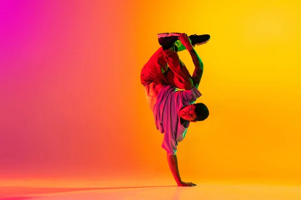 Plná délka portrét mladého stylového muže, break dance tanečnice v neformálním oblečení izolované přes gradient růžové žluté pozadí v tanečním sále v neonovém světle. — Stock fotografie