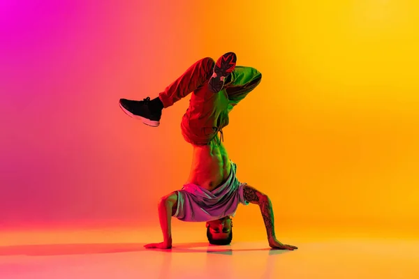 Portret van jonge stijlvolle man, breakdance danseres training in casual kleding geïsoleerd over hellend roze gele achtergrond in danszaal in neon licht. — Stockfoto